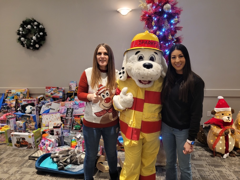Two volunteers with Sparky the fire dog beside tables of donated toys